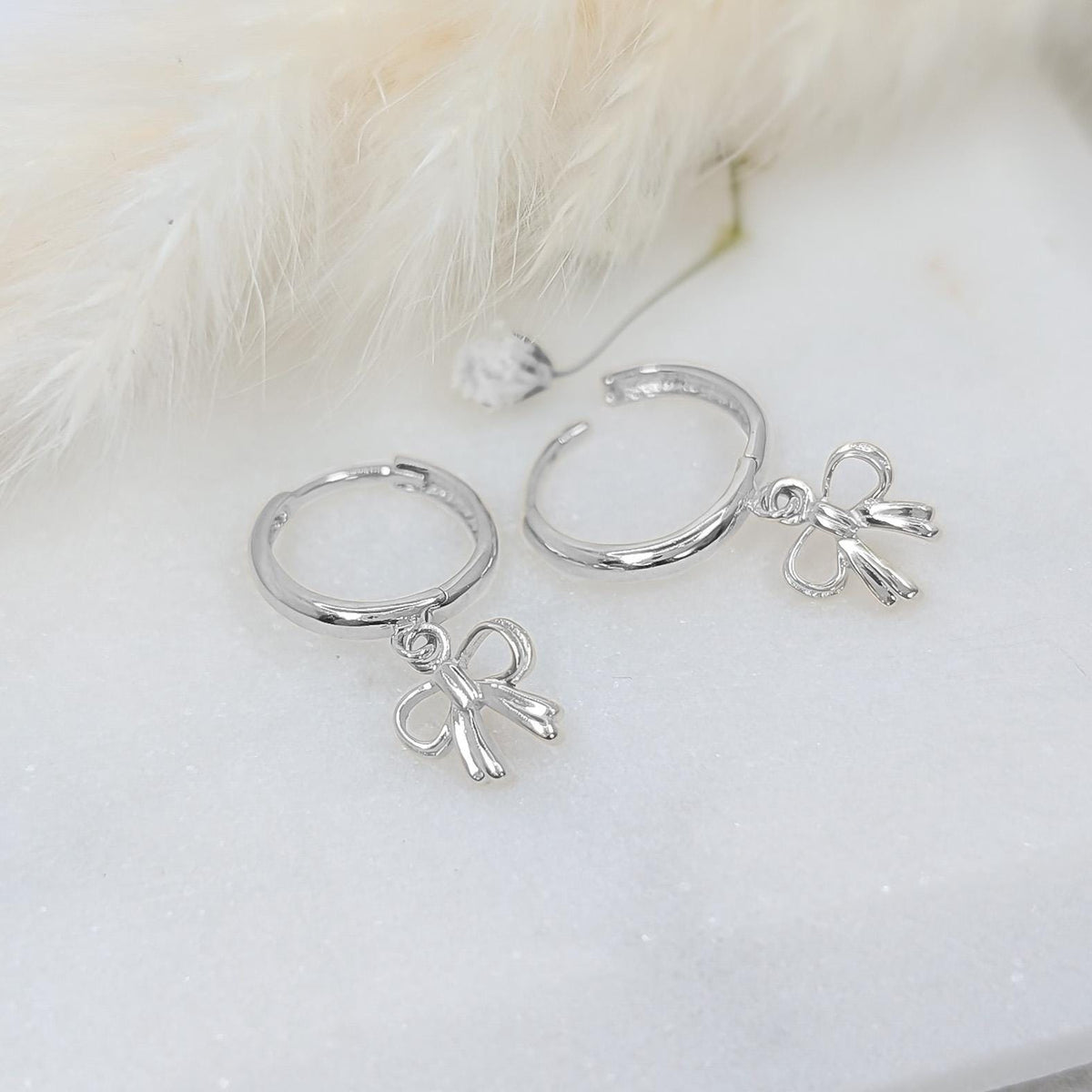 May include: A pair of silver hoop earrings with bow-shaped charms. The earrings are displayed on a white surface with a soft, feathery accent in the background. The bows are detailed and add a delicate touch to the jewelry.