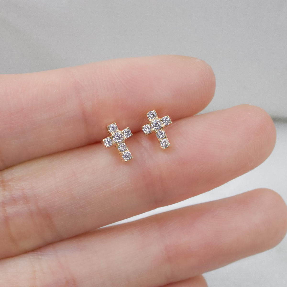May include: A pair of small, gold-colored cross stud earrings. Each cross is encrusted with small, clear gemstones. The earrings are displayed on a light-colored background.