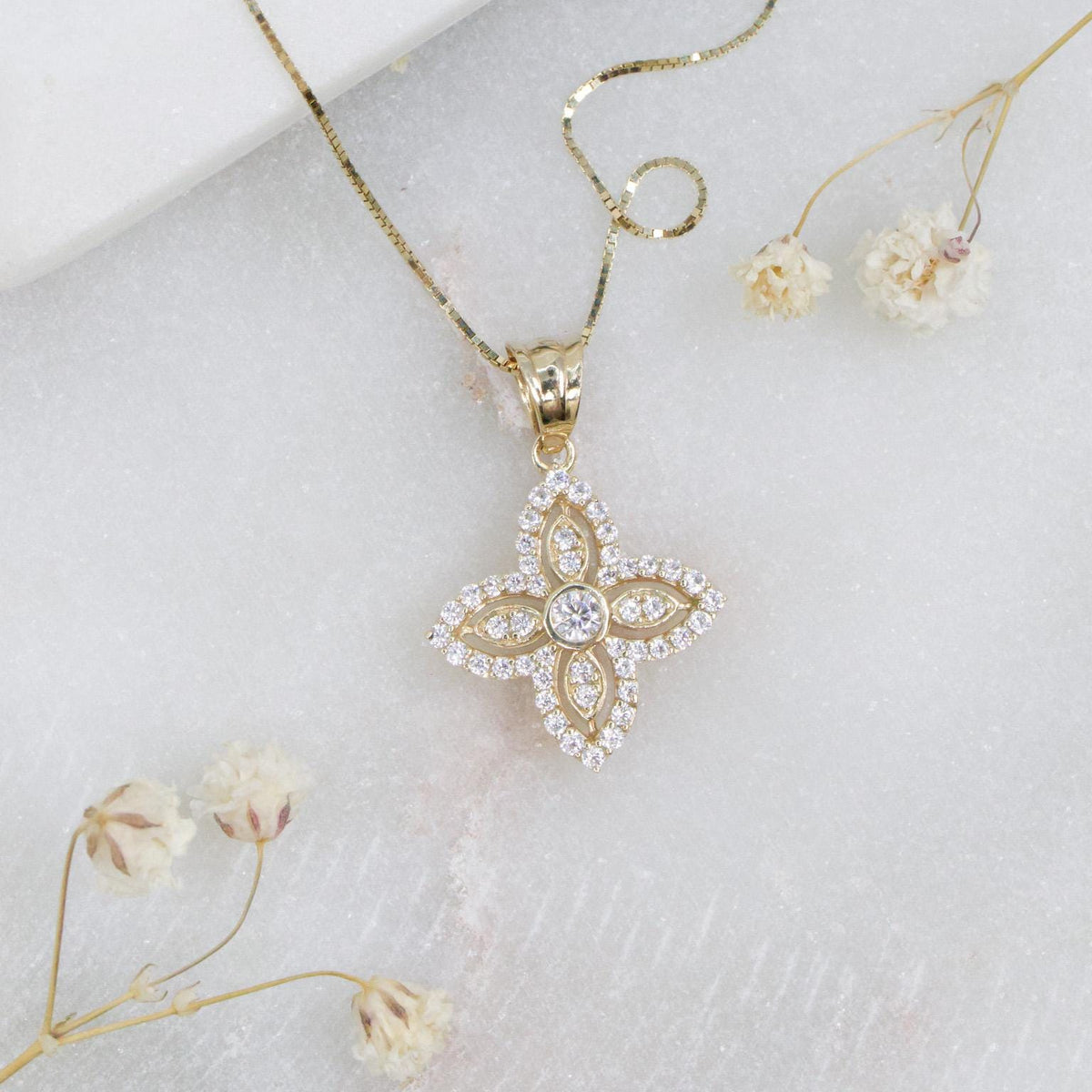May include: A gold-colored pendant necklace featuring a four-petal flower design. The pendant is encrusted with small, clear gemstones and a larger central stone. The necklace rests on a white surface, with dried flowers nearby.