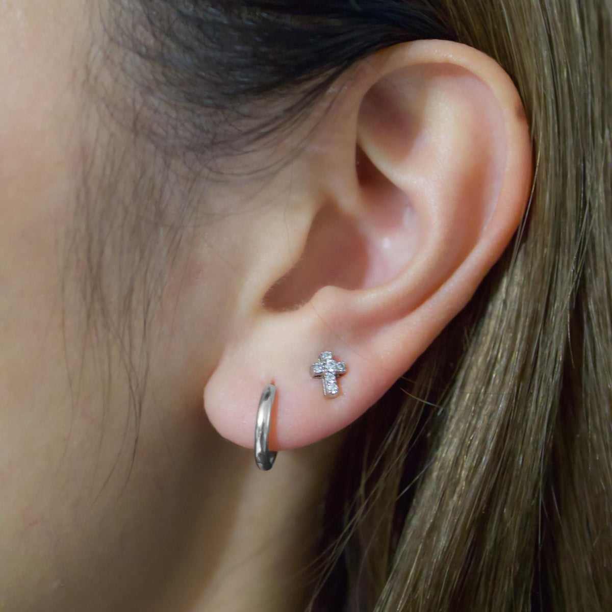 May include: A close-up of an ear with two earrings. One is a small silver hoop, and the other is a cross-shaped stud with small, clear stones. The ear is set against a blurred background of brown hair.